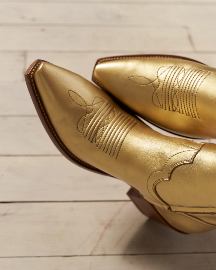 Golden cowboy boots with intricate stitching detail, resting on a light wooden floor.