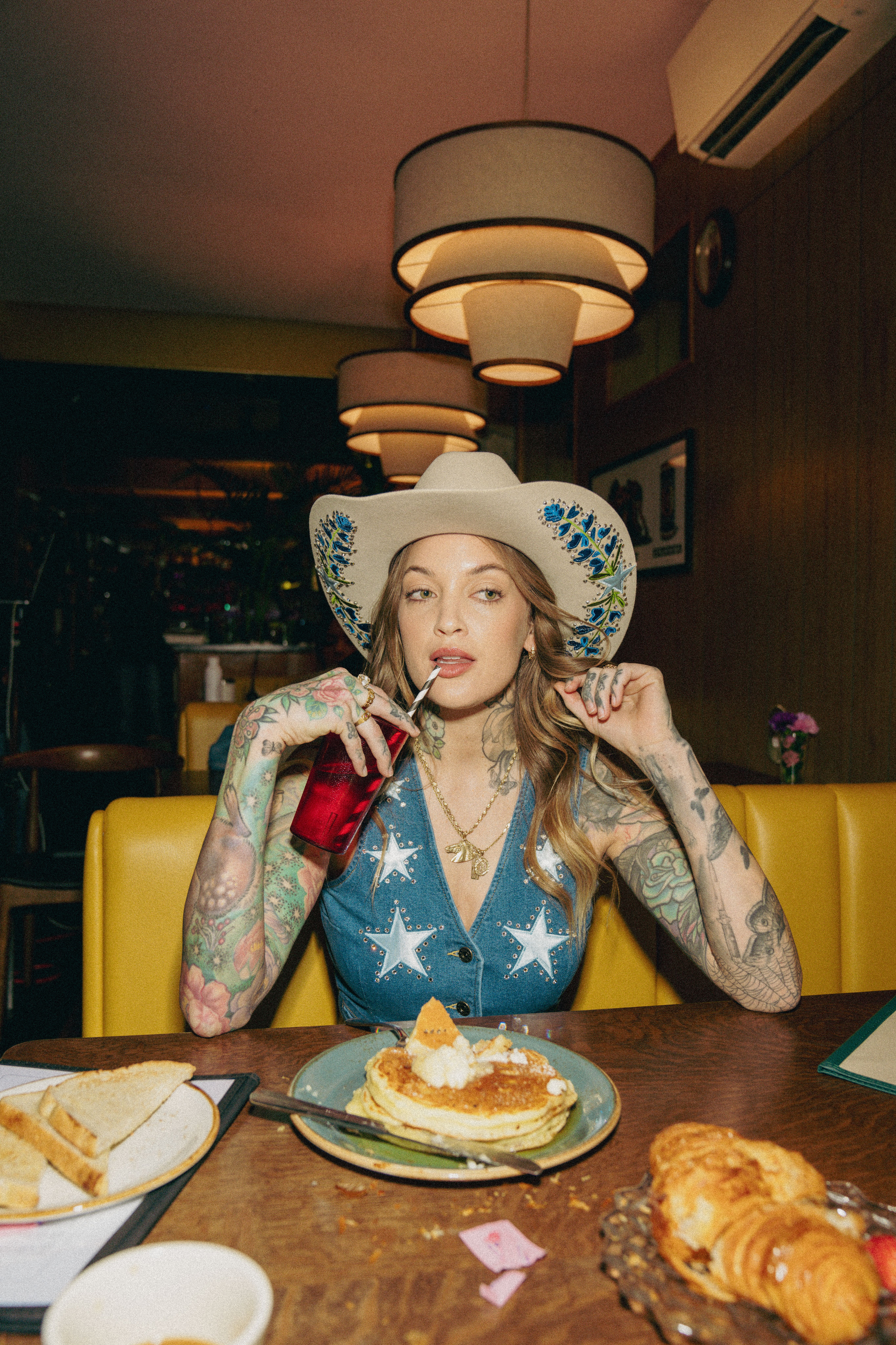 A person in a star-patterned outfit and hat drinks from a red glass with a straw at a diner table with pancakes and pastries.
