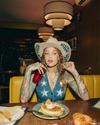 A person in a star-patterned outfit and hat drinks from a red glass with a straw at a diner table with pancakes and pastries.