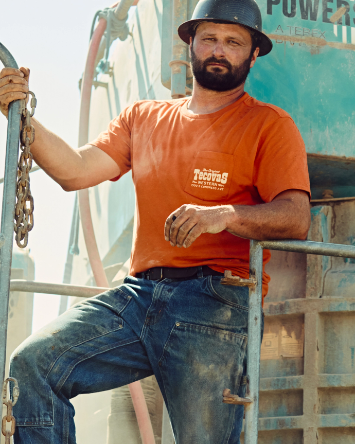 A man wearing a black hard hat, orange Tecovas t-shirt, and jeans stands on metal steps by industrial equipment, holding a chain with one hand.