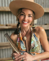 A smiling person wearing a wide-brimmed hat and colorful plaid top holds a feather. They are accessorized with rings, a bolo tie, and braids, standing against a corrugated metal backdrop.