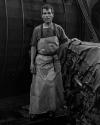A man wearing a wet apron stands beside a large pile of animal hides inside an industrial setting, with metal machinery in the background.