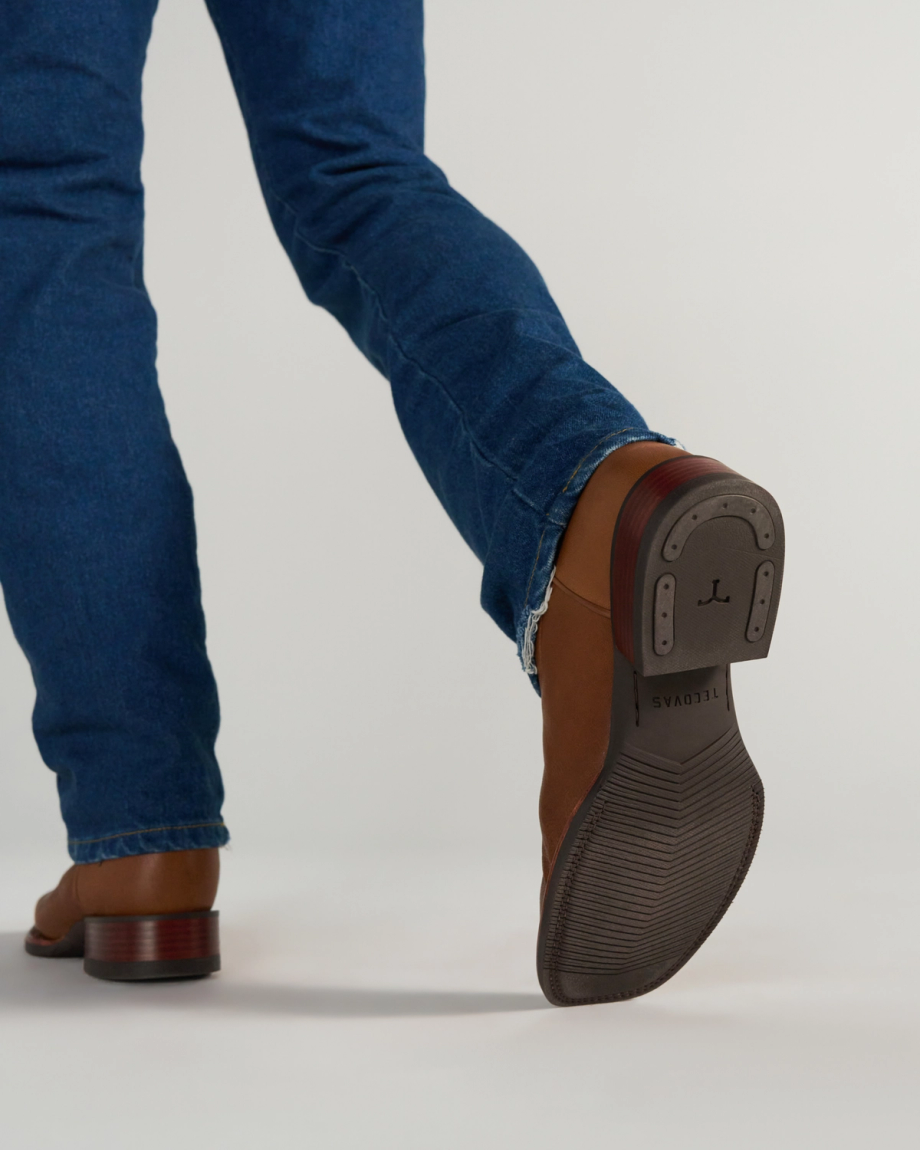 The Dusty in Mesa Goat. Person wearing blue jeans and brown heeled boots, lifting one foot to show the sole with a metal plate and engraved markings, against a plain background—an understated nod to Mesa Goat style.