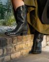 Close-up of a person standing on a brick curb, wearing textured black cowboy boots and a green trench coat with a black bag.