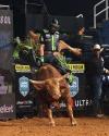 A bull rider balances on a bucking bull in an arena, wearing protective gear and surrounded by dirt and arena signage.