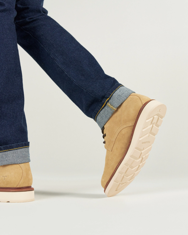 A person wearing cuffed dark denim jeans and tan The Knox Lace Up Shearling Roughout shoes in Sandstone Roughout stands on a white surface, with one foot slightly raised to show the rugged shoe sole.
