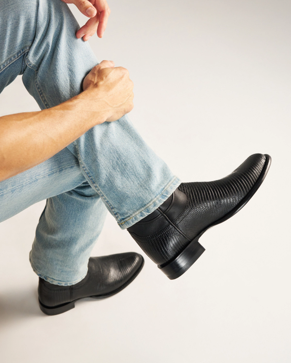 The Earl in Obsidian Lizard. A person wearing light blue jeans and textured black leather Obsidian Lizard boots sits with one leg crossed over the other, holding their ankle.