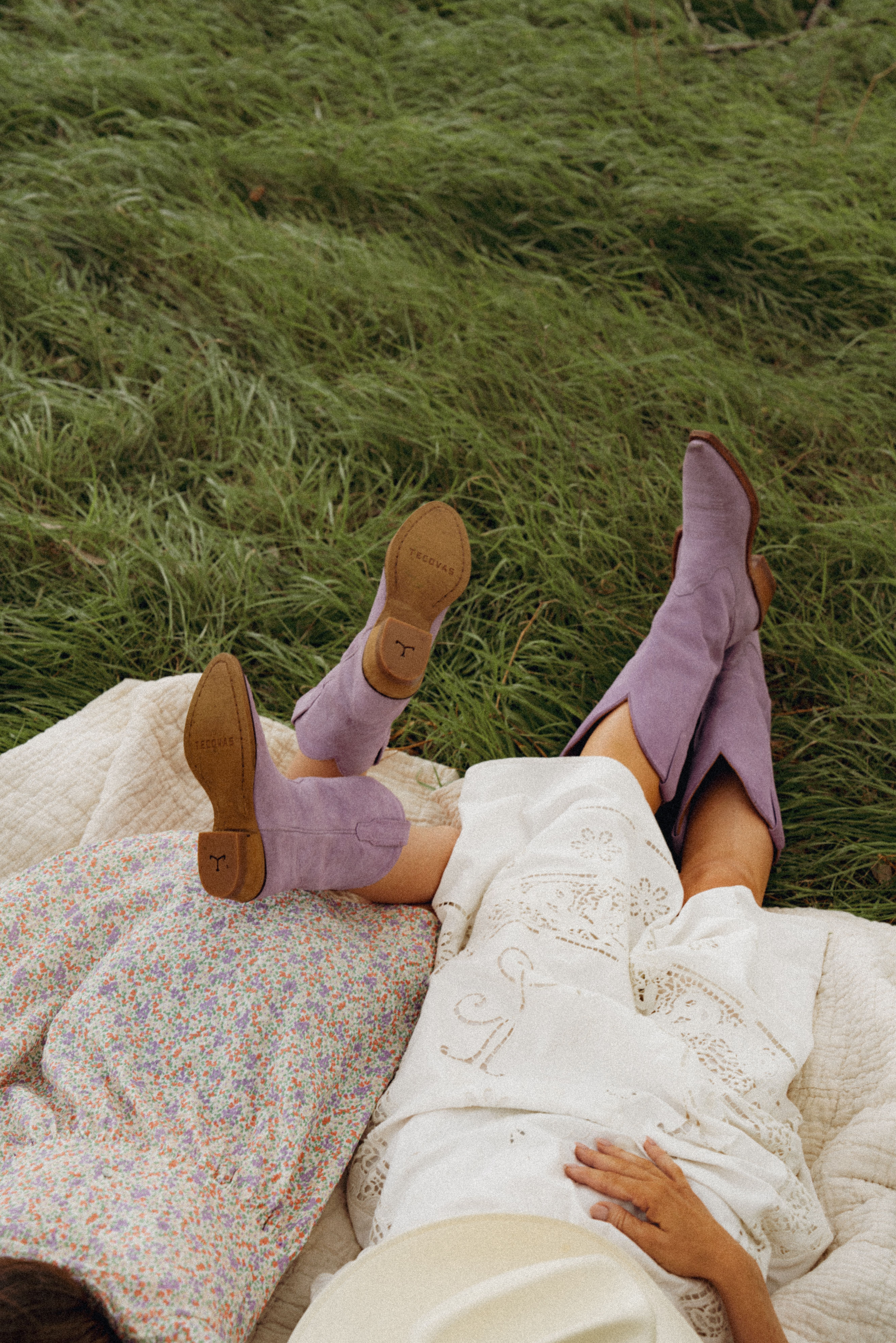 Two people lie on a blanket on grass; one wears a floral dress, the other a white embroidered dress and purple boots, both with legs outstretched.