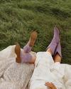 Two people lie on a blanket on grass; one wears a floral dress, the other a white embroidered dress and purple boots, both with legs outstretched.