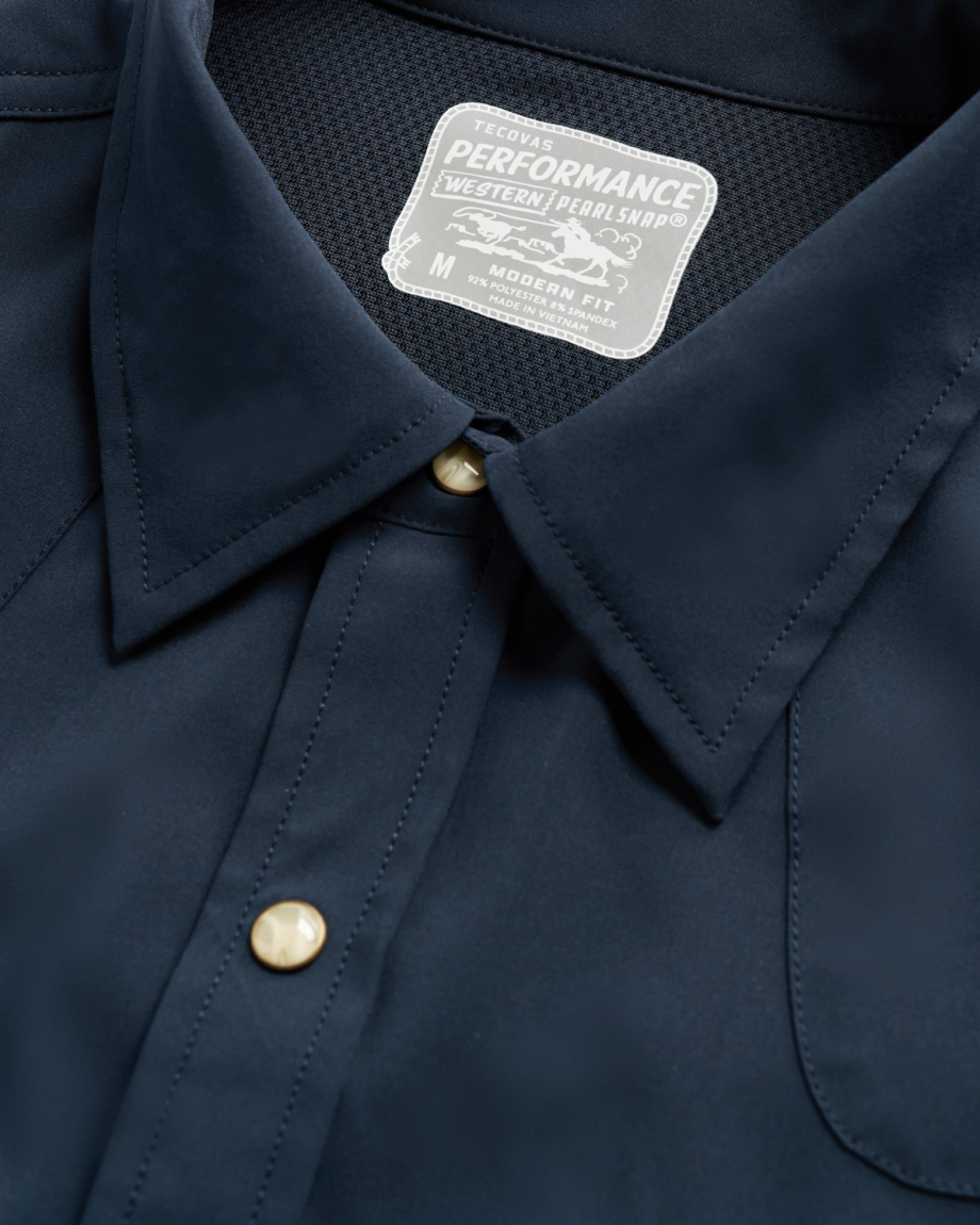 Close-up of a navy blue shirt with gold snap buttons and a white label that reads "Tecovas Performance Western Pearl Snap, Modern Fit, M.