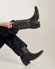 Person wearing dark denim jeans and black embroidered cowboy boots, holding one boot by the side against a plain background.