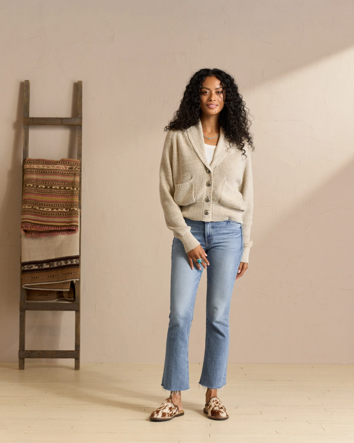 A person with long curly hair wearing a beige cardigan, white top, blue jeans, and patterned shoes stands in a room with a ladder and folded blankets.