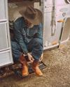 A person wearing a hat and denim outfit sits on the step of an RV, putting on brown boots.