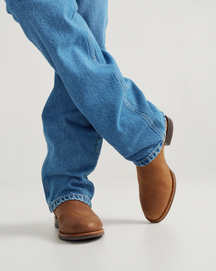 The Glen in Khaki Cowhide. Person wearing light blue jeans and The Glen Cowhide brown leather boots stands with legs crossed against a plain background.
