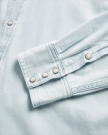 Close-up of a light blue denim shirt sleeve and cuff, featuring metal snap buttons and visible stitching details.