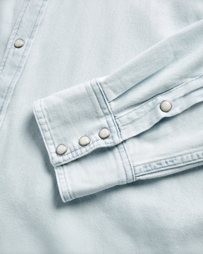 Close-up of a light blue denim shirt sleeve and cuff, featuring metal snap buttons and visible stitching details.