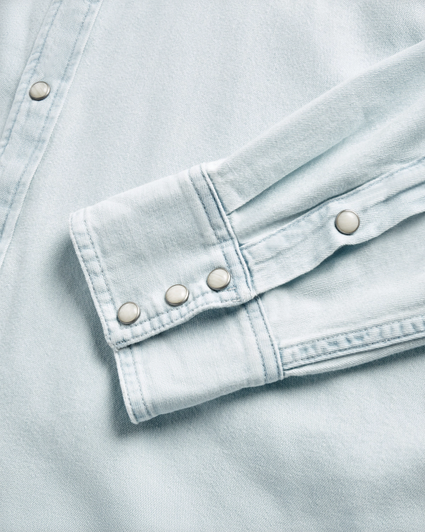 Close-up of a light blue denim shirt sleeve and cuff, featuring metal snap buttons and visible stitching details.