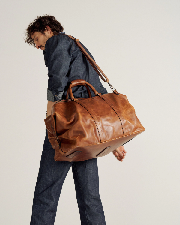 Person in a denim outfit carrying a large brown leather duffel bag over one shoulder, photographed against a plain background.