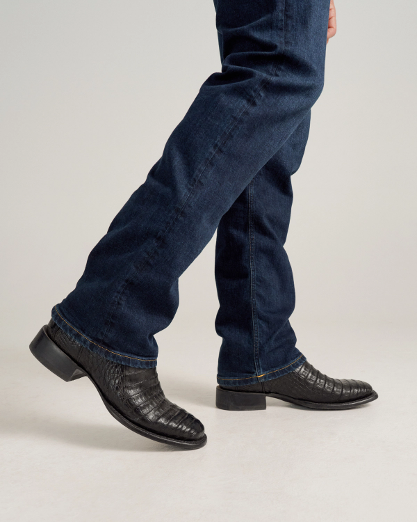 A person wearing midnight blue jeans and The William Caiman black crocodile-patterned dress shoes is walking on a light surface.