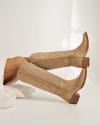 A person lying down wears tan suede knee-high cowboy boots with white stitching and a low wooden heel, set against a plain white background.