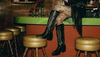 Tattooed person sitting on a bar counter, wearing black cowboy boots and a fringed outfit, next to gold stools, with roses in the background.