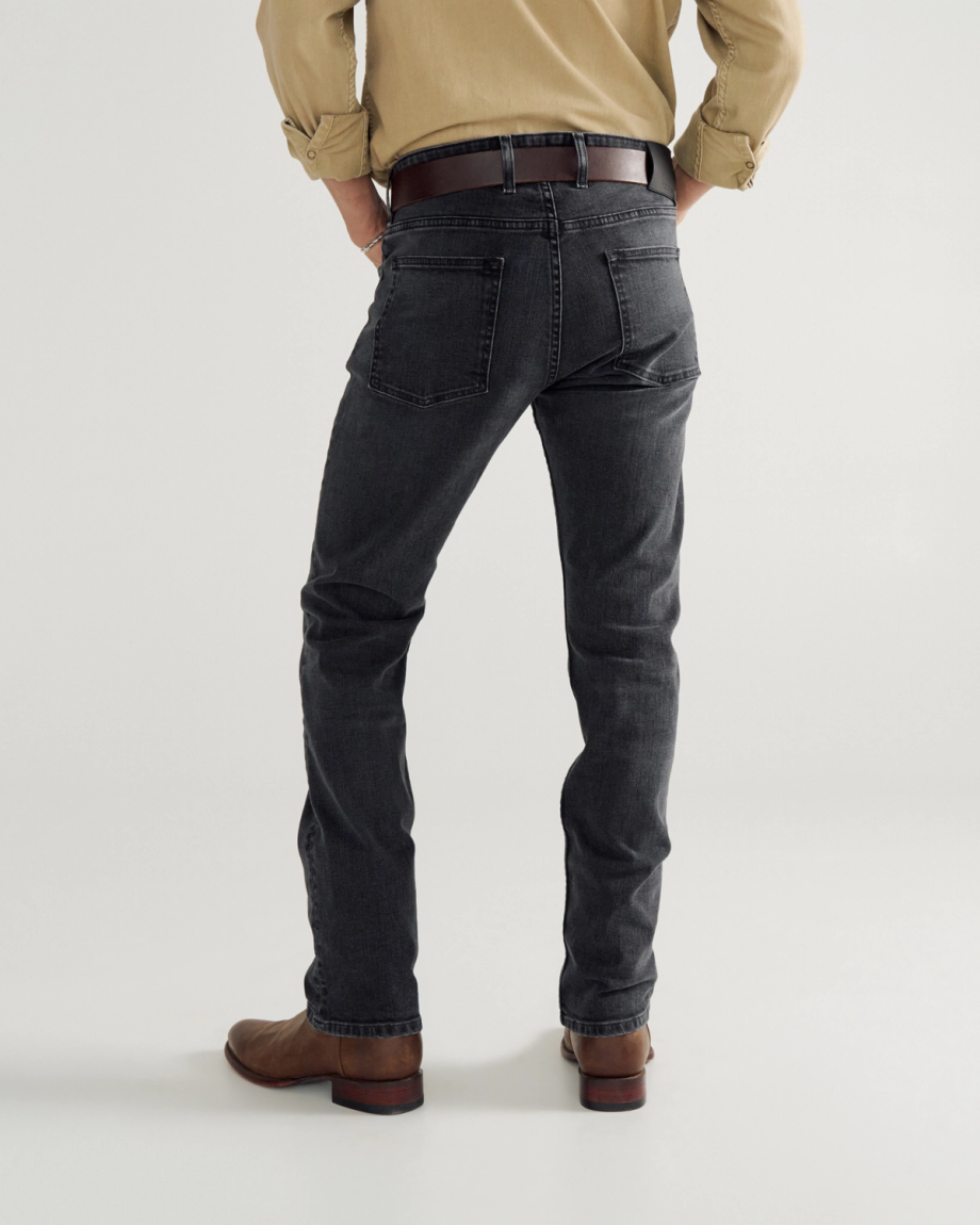 A person wearing a tan shirt, black jeans with a brown belt, and brown boots stands facing away from the camera against a plain background.