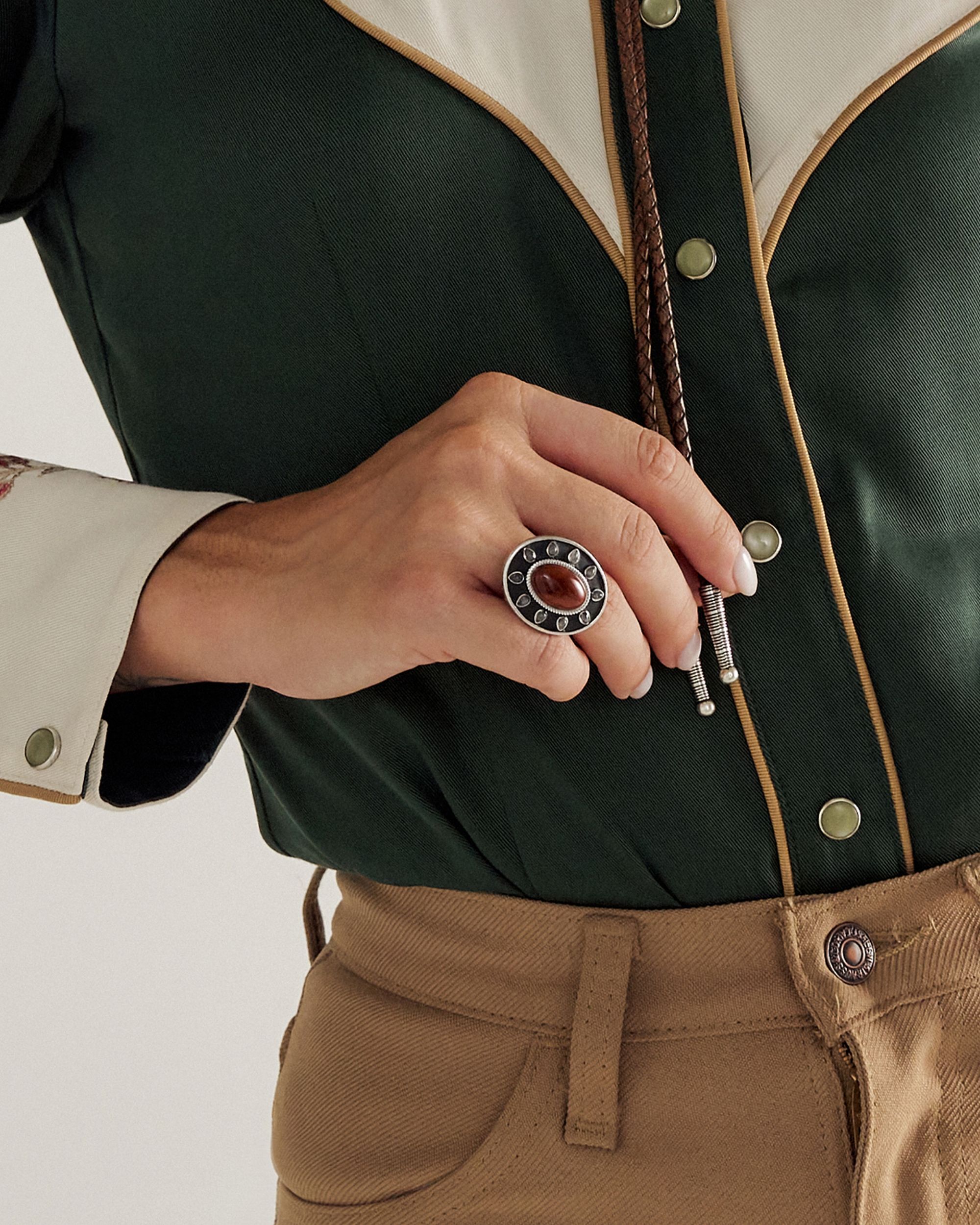 A person wearing a green shirt with embroidered cuffs and tan pants, holding a bolo tie and wearing a large decorative ring on their finger.