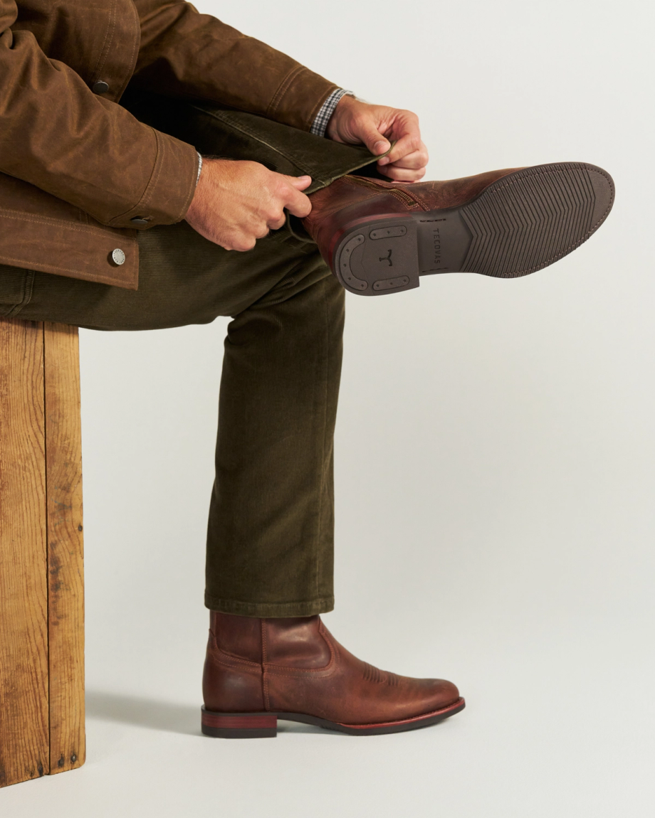 The Wayne in Maplewood Cowhide. A person sits on a maplewood bench, wearing brown pants and a brown jacket, putting on a classic Wayne Cowhide leather boot.