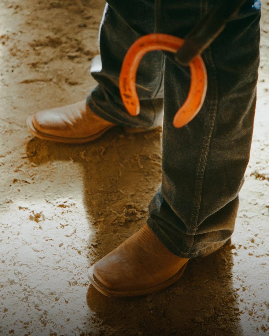 Person wearing jeans and brown boots standing on dirt, holding a horseshoe in one hand.