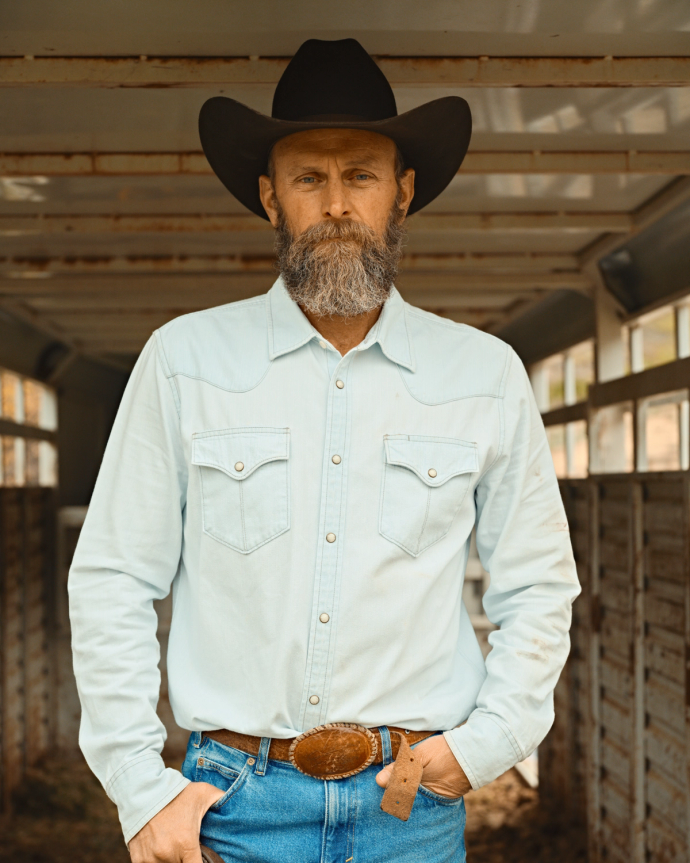 A man with a beard and mustache wears a black cowboy hat, light blue shirt, and blue jeans, standing confidently inside a rustic building.