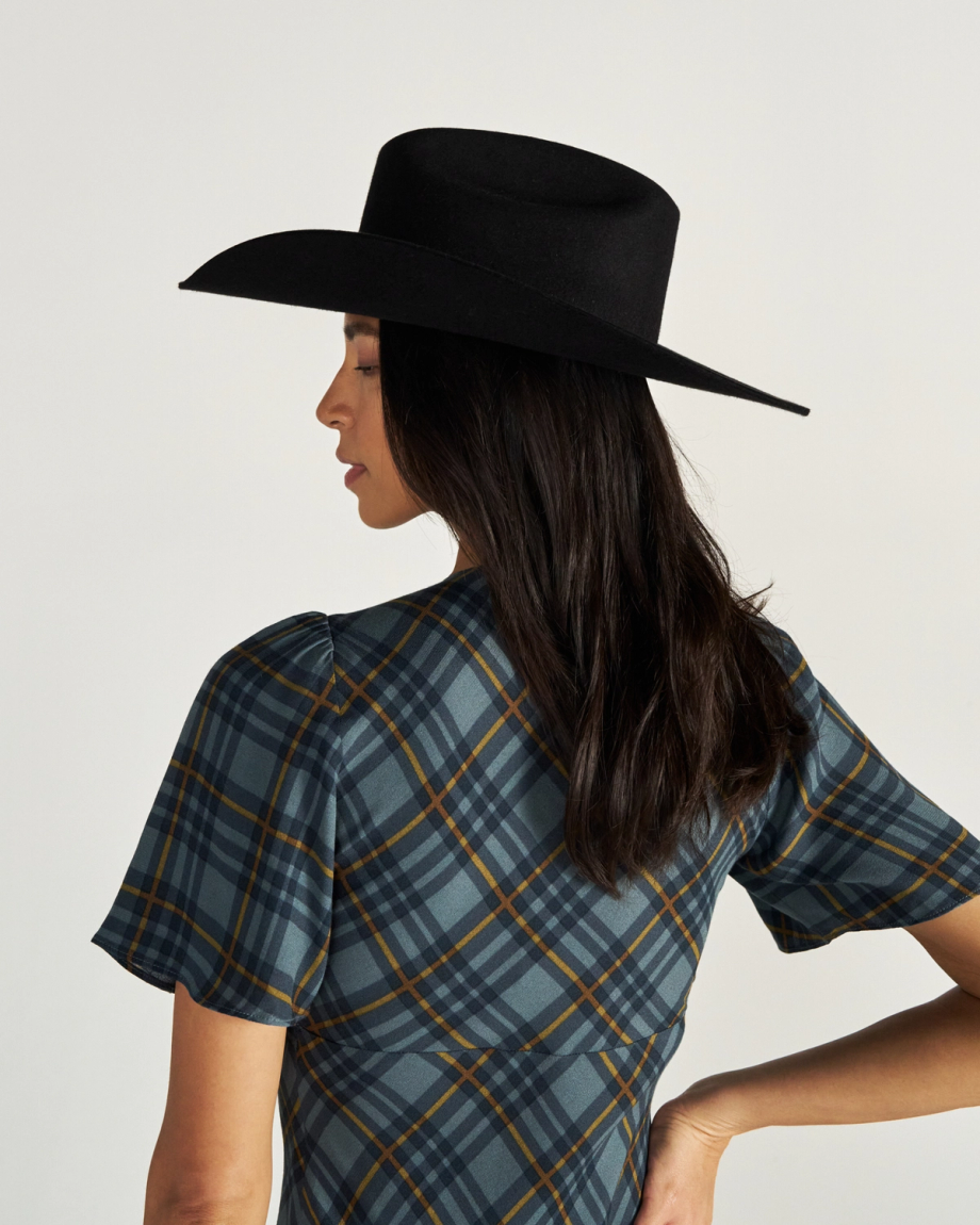Woman in a plaid dress and black wide-brim hat stands facing away, showing her profile and long dark hair against a plain background.
