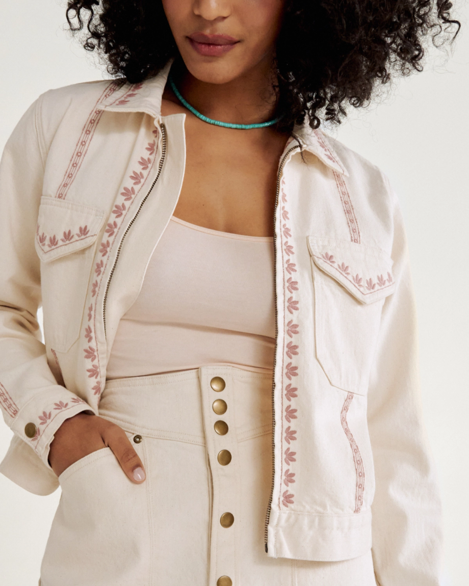 Person wearing a light-colored jacket with pink embroidery, matched with a beige top and skirt featuring gold buttons. They are also wearing a turquoise necklace Natural Denim .