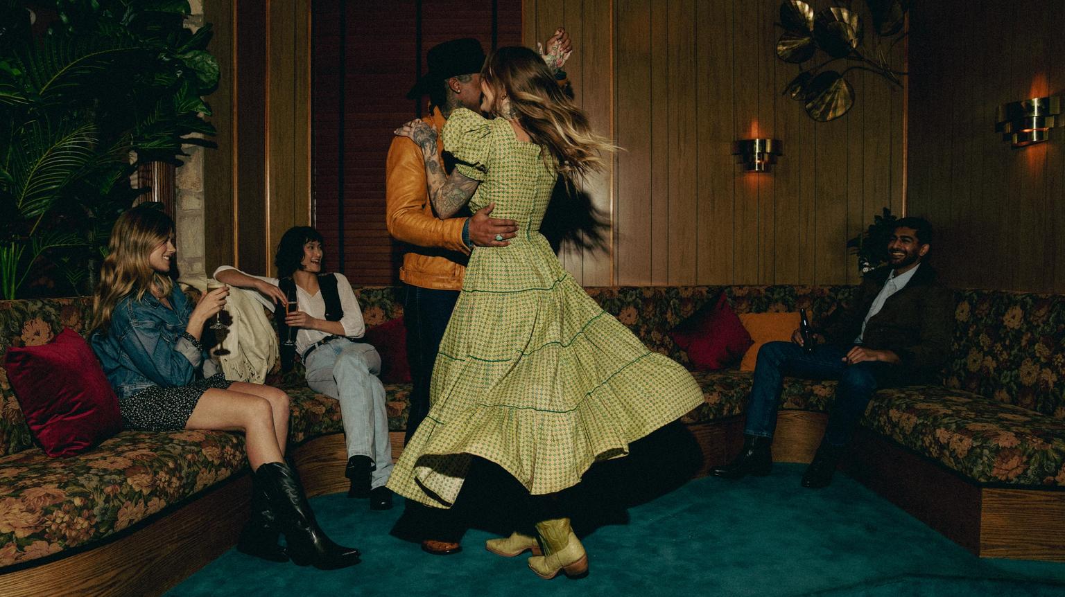 A couple dances joyfully in a dimly lit room while three people seated on a floral couch watch, next to a wood-paneled wall with plants and decorative lighting.