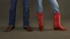 Two people stand side by side, one wearing brown leather cowboy boots and blue jeans, the other wearing red embroidered cowboy boots and blue jeans, against a plain dark background.