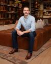 A man with a mustache, wearing a light blue shirt and dark jeans, sits on a brown leather bench in a store with shelves displaying boots and bags.