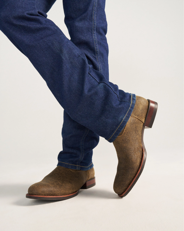 The Glen in Dustwood Roughout. Person wearing blue jeans and brown Dustwood suede boots stands with legs crossed against a plain background.