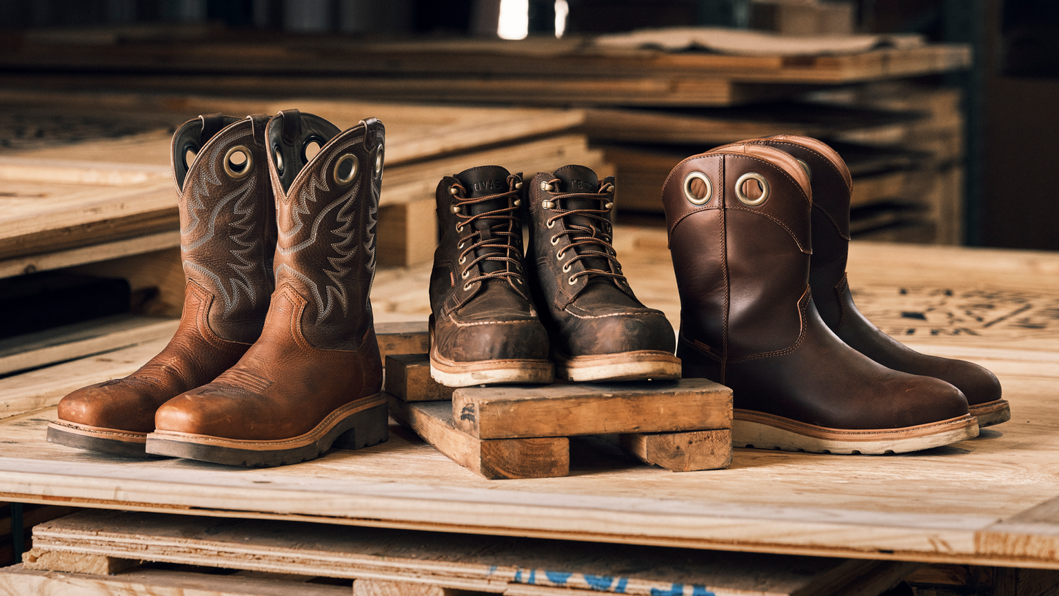 Three pairs of brown work boots, each with distinct designs, are displayed on wooden pallets and boards.