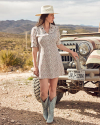 A woman in a floral dress and cowboy boots stands beside an old jeep in a desert landscape, wearing a wide-brimmed hat.