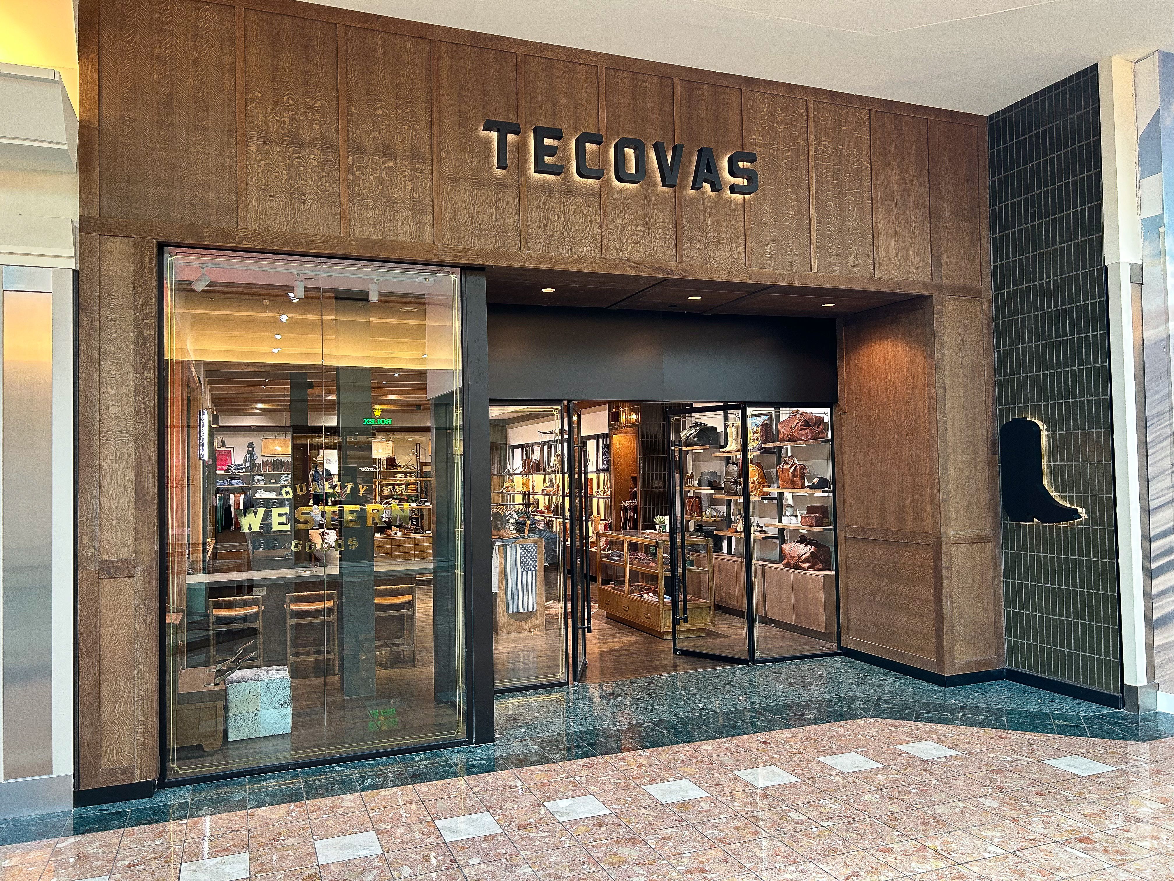 The entrance to a Tecovas store in a mall, featuring wood paneling, glass doors, and displays of boots and leather goods inside.