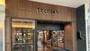 The entrance to a Tecovas store in a mall, featuring wood paneling, glass doors, and displays of boots and leather goods inside.
