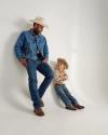 A man and a young boy, both wearing cowboy hats, jeans, and boots, stand and sit against a plain white wall, looking at each other.