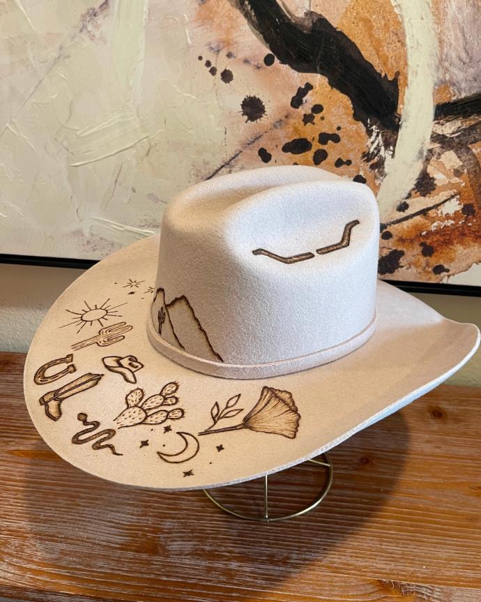 A light tan cowboy hat with brown embroidered designs, including a sun, snake, cactus, moon, and mountains, displayed on a wooden surface in front of an abstract painting.