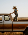 A person wearing a cowboy hat and denim outfit stands in the bed of an old pickup truck parked in a dry, open field.