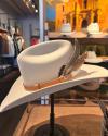 A cream-colored cowboy hat with a brown band and decorative feathers displayed on a stand in a store.
