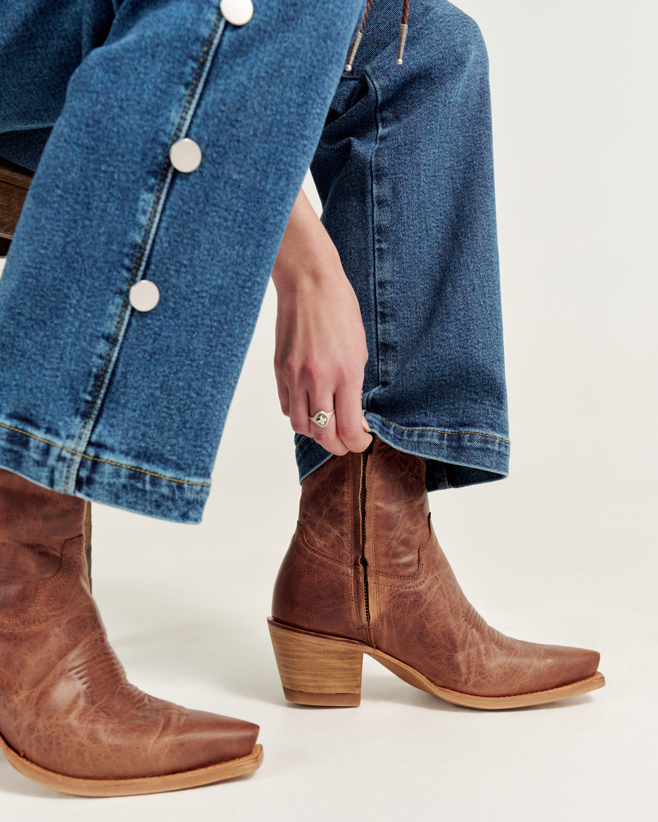 Person wearing blue jeans with white buttons, adjusting a brown leather cowboy boot with a block heel.