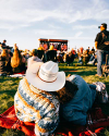 Couple sitting on a blanket in a park, watching a concert. The person wearing a cowboy hat leans against the other, surrounded by a crowd.