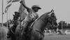 Black and white photo of a man riding a horse with an american flag. Keywords: Photo, horse, American flag.