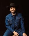 A person wearing a black cowboy hat and blue denim jacket sits smiling against a dark background.