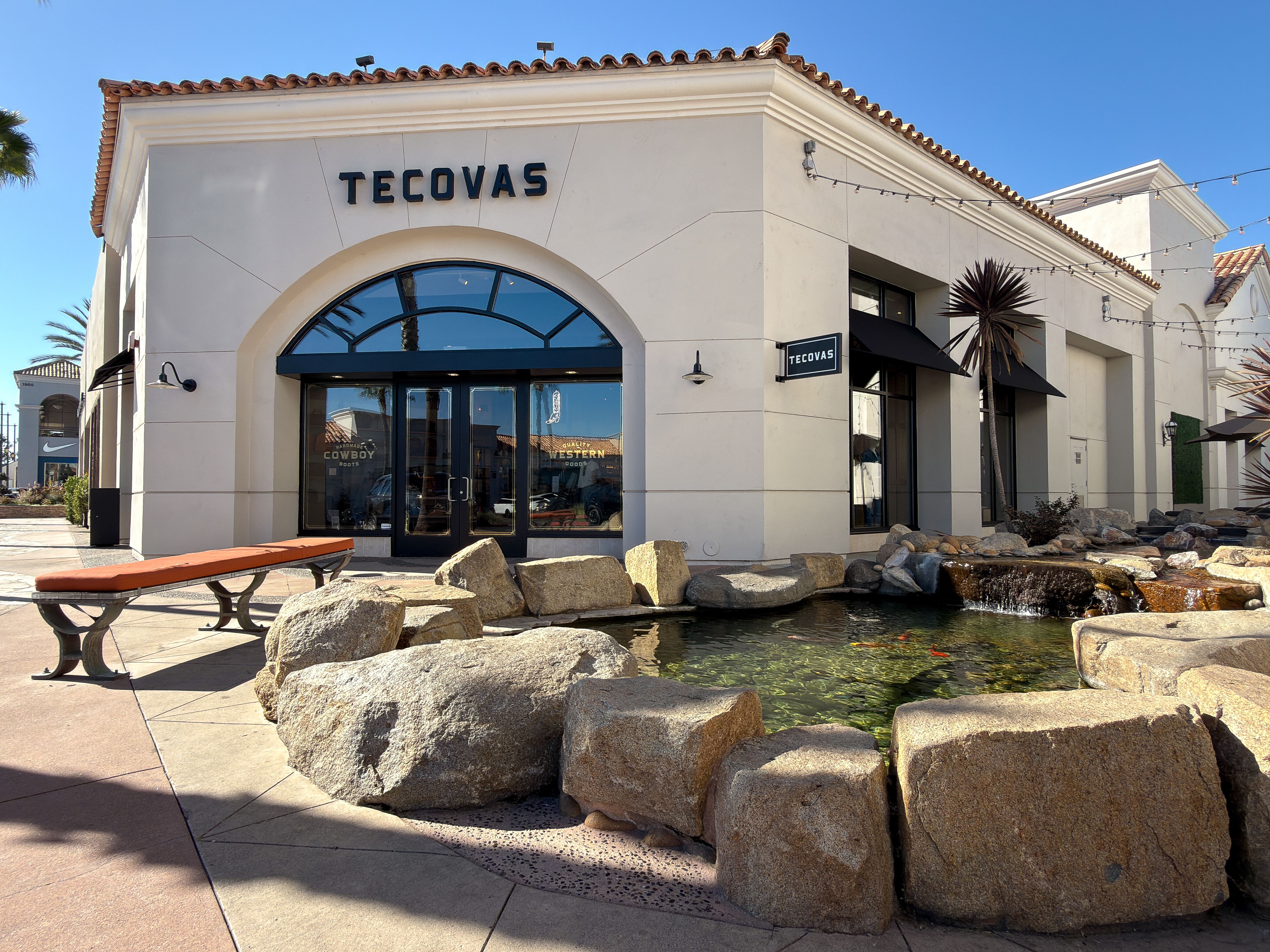 A Tecovas storefront with arched glass doors, a rock-lined pond in front, a wooden bench, and clear sunny skies.
