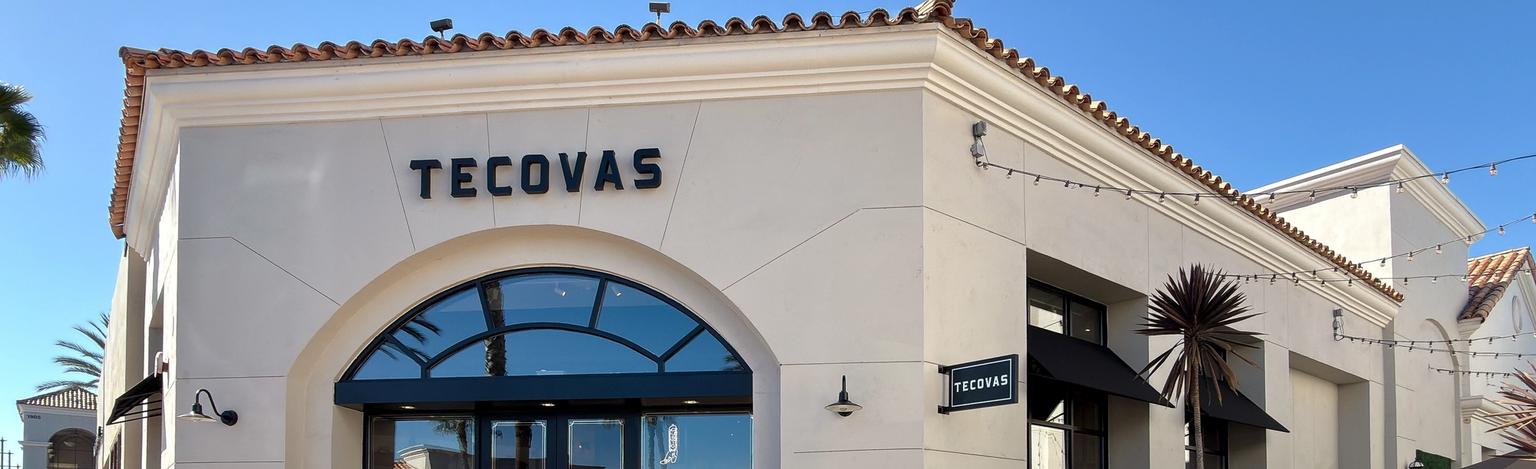 A Tecovas storefront with arched glass doors, a rock-lined pond in front, a wooden bench, and clear sunny skies.